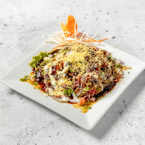 A colorful Indian chaat dish, topped with sauces, spices, and crispy noodles, on a white rectangular plate.