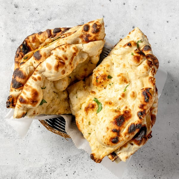 A basket of freshly baked, golden-brown naan bread with charred spots and green herbs.