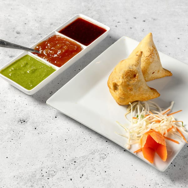 Two golden-brown samosas with shredded cabbage and carrot garnish, served with three dipping sauces.