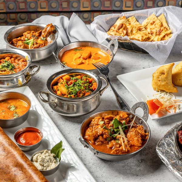 A table laden with various Indian dishes in metal bowls, including curries, naan, and samosas.