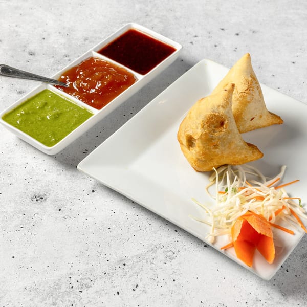 Two golden-brown Nirvana Indian Kitchen samosas with shredded vegetables, and a trio of dipping sauces.