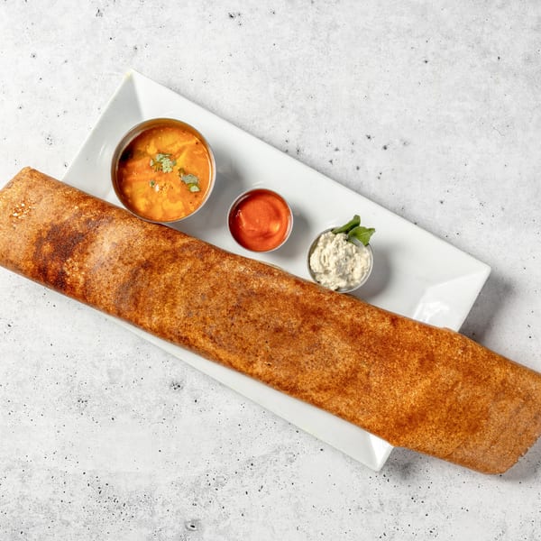 A long, crispy dosa with three small bowls of Indian chutneys and sambar on a white plate.