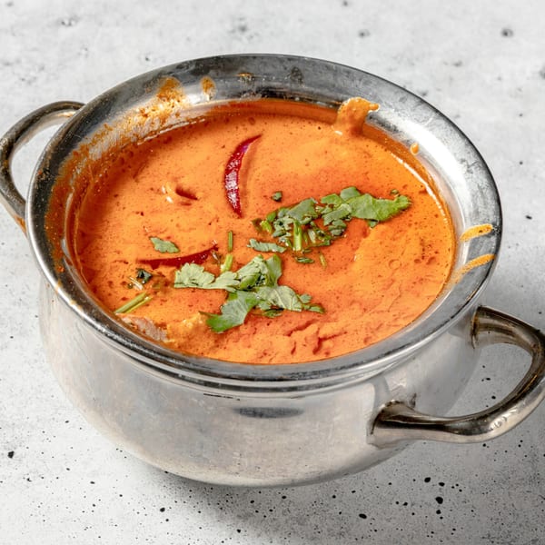 Vibrant orange Indian curry, garnished with fresh cilantro and a red chili, in a silver serving pot.